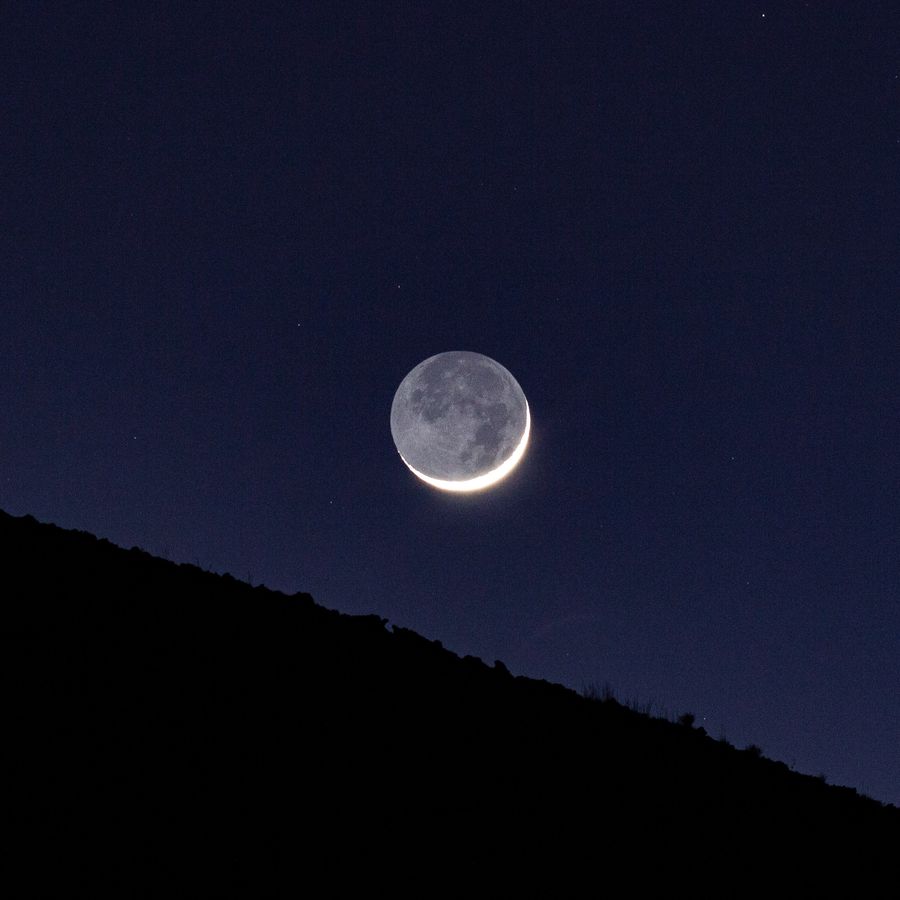A crescent Moon with earthshine setting behind a ridge